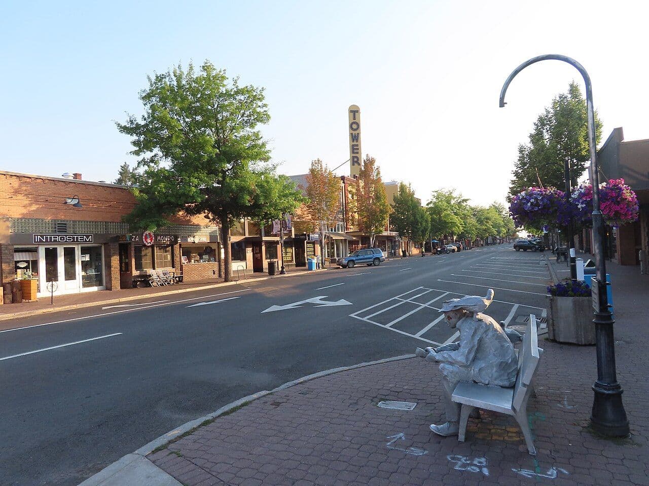 A downtown Bend street with the Tower Theatre sign, hanging flower baskets and a bronze bench statue.